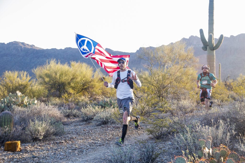 man with peace sign american flag running on sandy dirt through cacti and shrubs