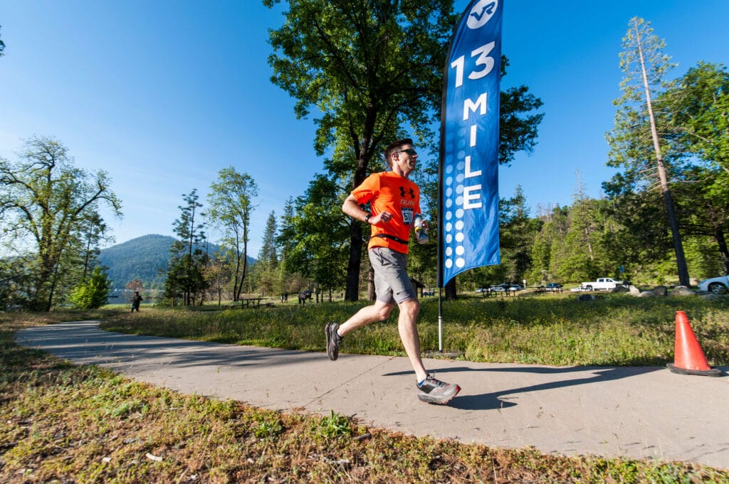 Adult running past a flag that says Mile 13 with trees and a lake behind him