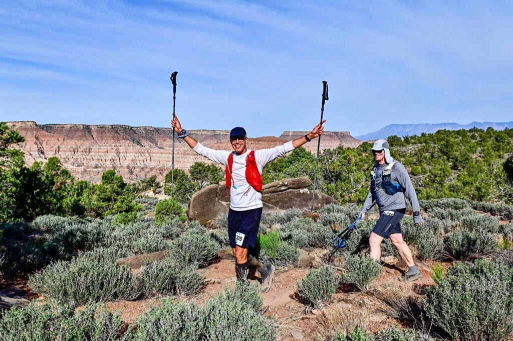 two runners walking along the mesa ledge. One has their trekking poles raised in the air.