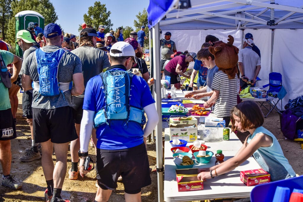 A busy aid station with volunteers passing out fruit, cookies, and other snacks as runners walk by