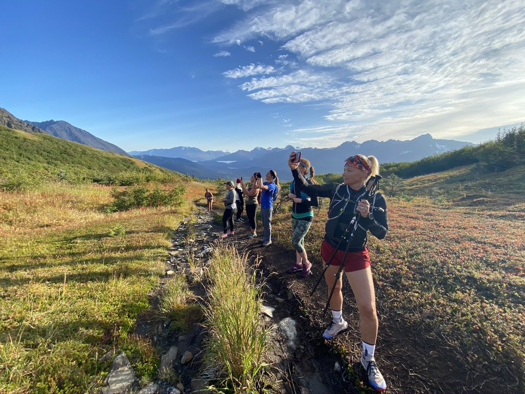A row of runners stop and take photos in a sunny grass field in between mountain passes near Mount Marathon