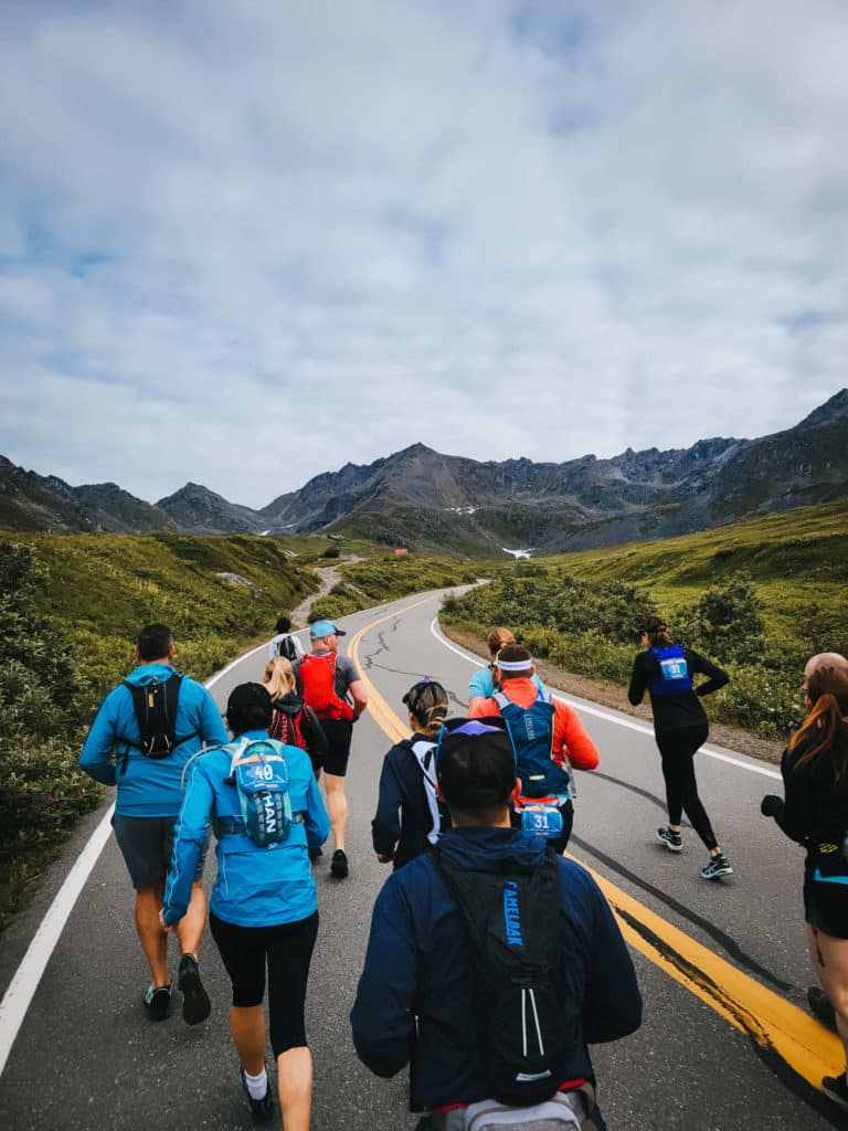 A group of runners in bright gear run along a wide empty road through a valley with grassy peaks and slopes