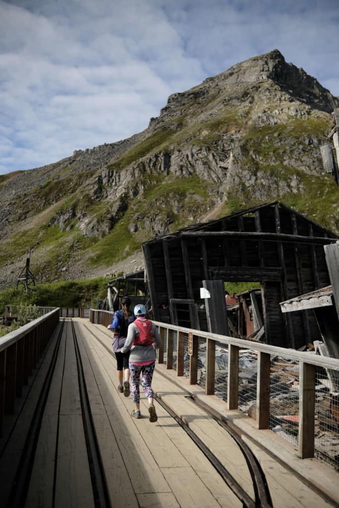 Two runners crossing an old wooden bridge with ruins from the pioneer days and a green slope rising in front of them