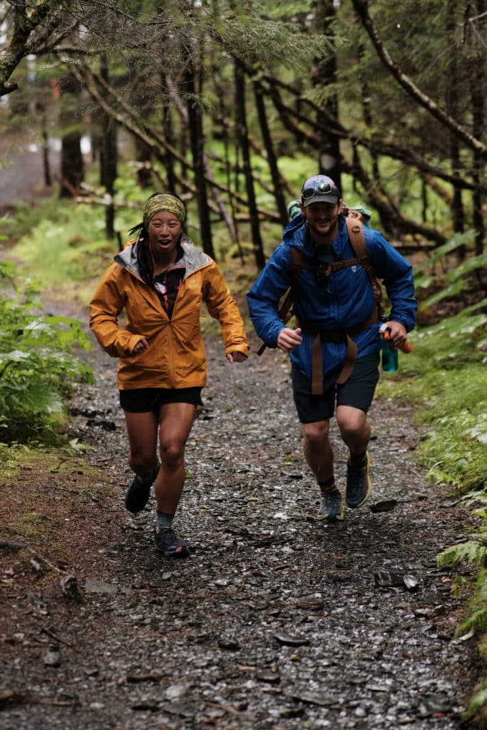 Two runners in rain gear ascend a dirt trail through a mossy forest