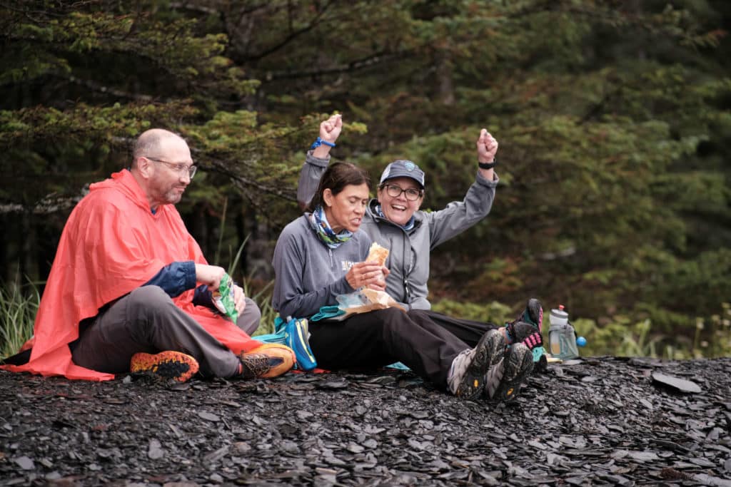 Three runners sit on the ground and eat a quick snack. One holds his hands up in victory after a hard run.