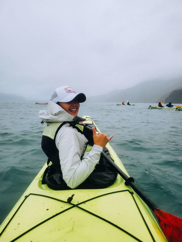 Young woman in a lime green ocean kayak smiling at the camera and holding up a 'rock on' hand gesture as she paddles into the fog