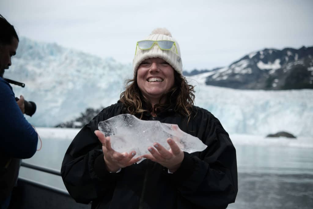 A smiling brunette woman holds up a block of ice while standing in front of a glacier