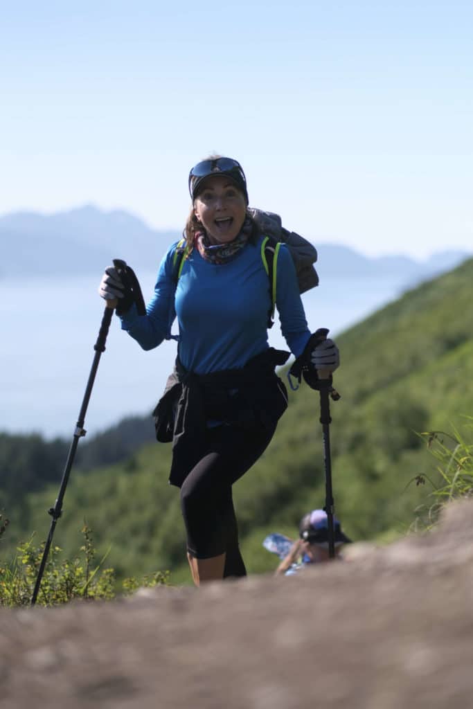Hiker coming over a hill with a big smile and trekking poles, behind her is water and mountains