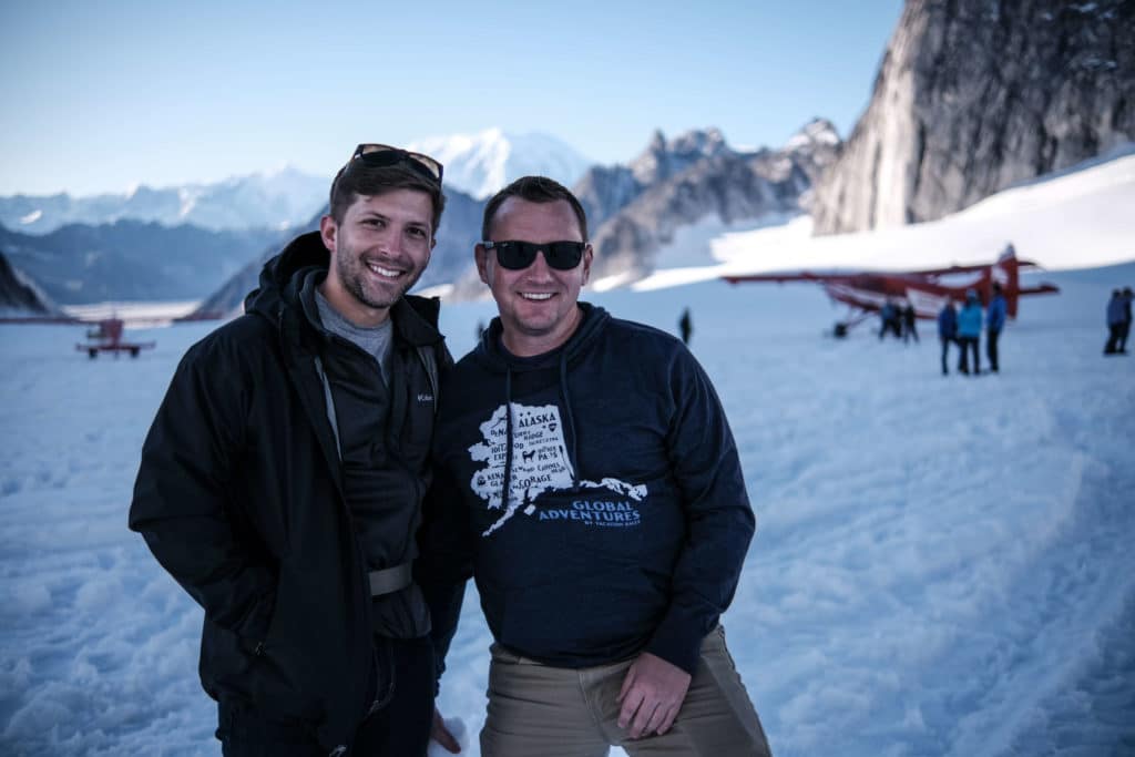 Two men smiling in winter gear with a pass of snow and mountains behind them in the distance