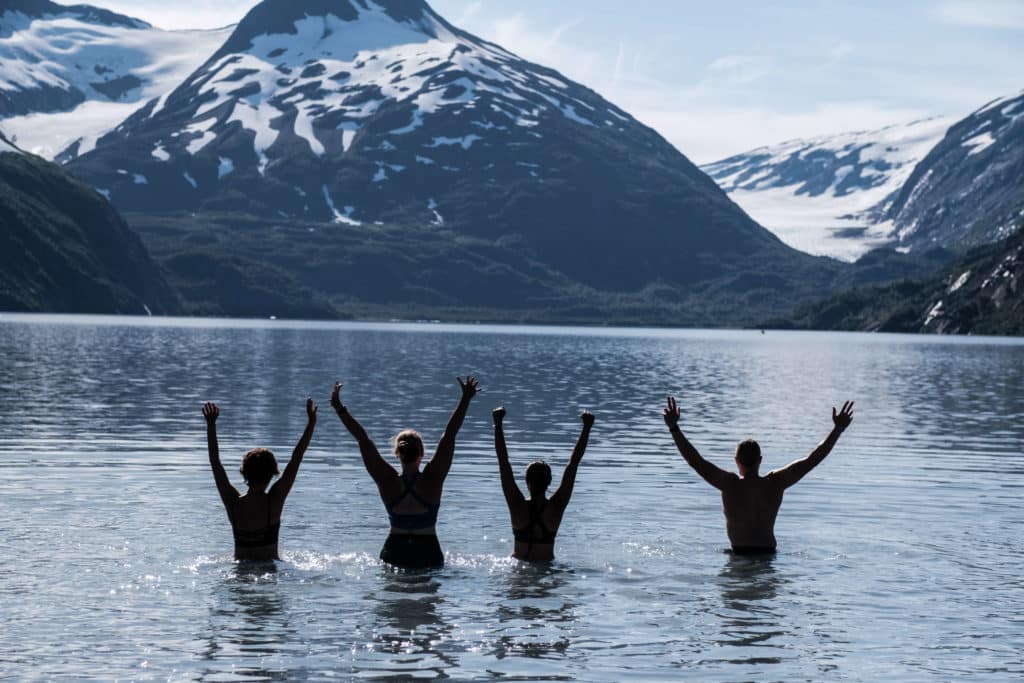 Four runners raise their arms as they wade into a mountain lake surrounded by peaks