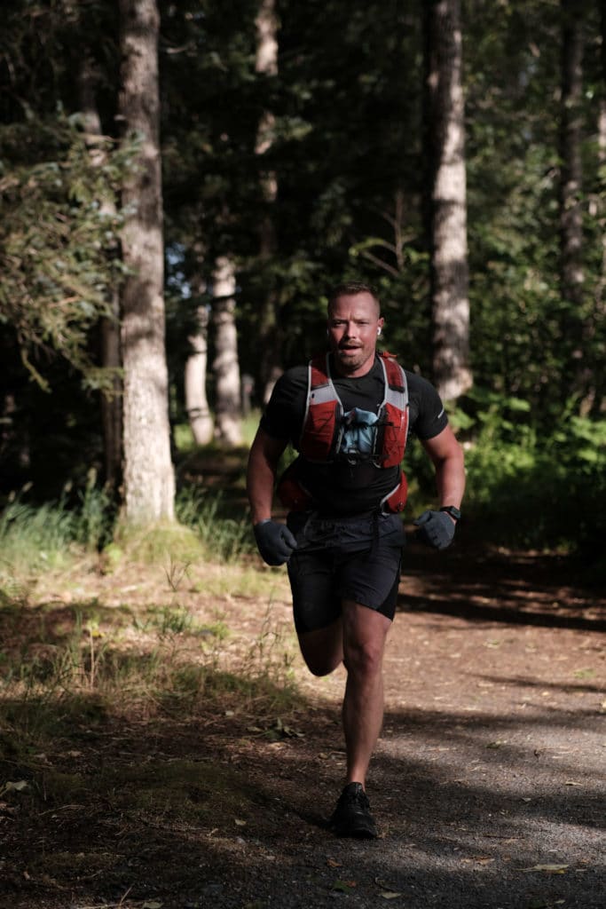 A man in a running vest runs fast through a sunny patch in a pine forest