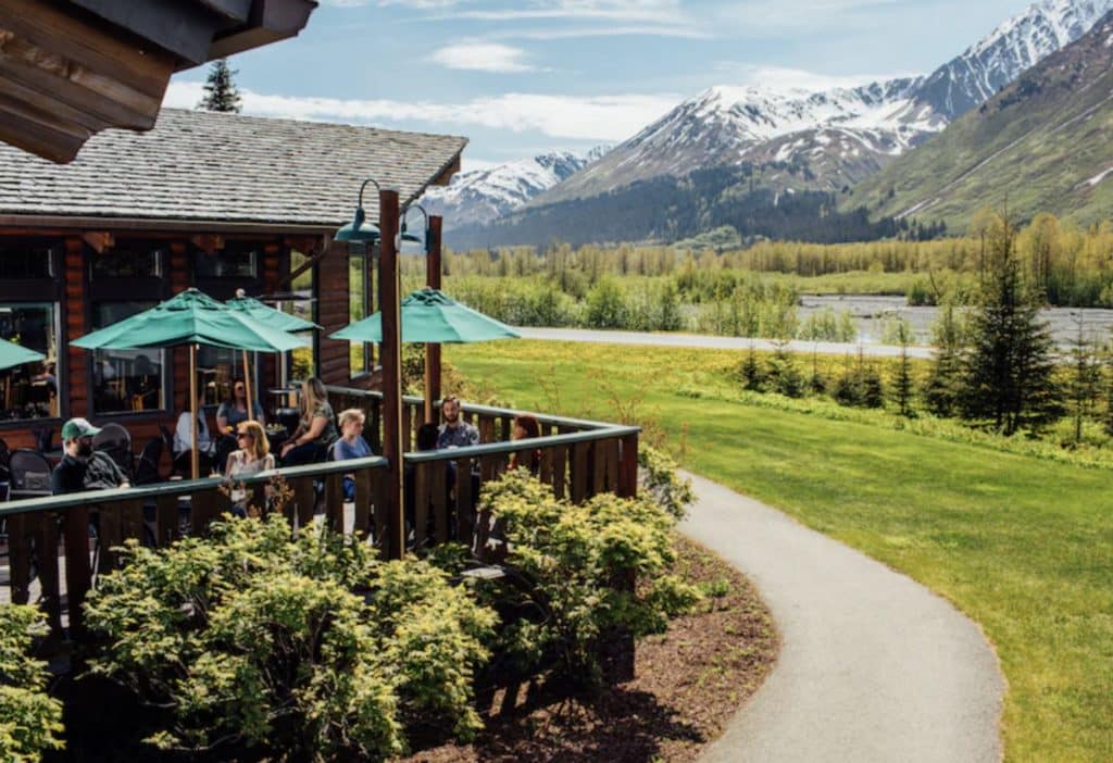 An outdoor dining deck at the Windsong Lodge with meadows and snow peaked mountains behind it
