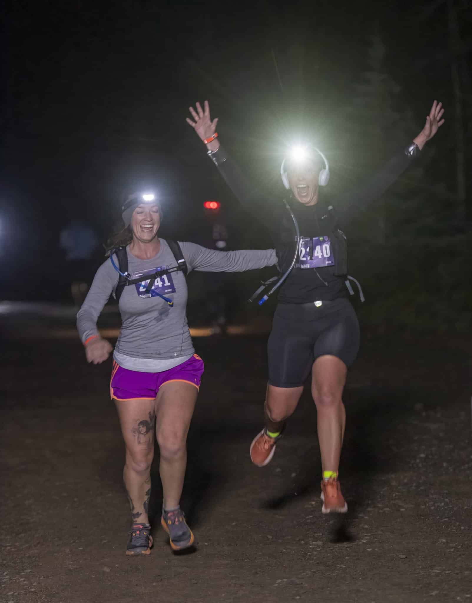 Two runners jump for joy as they approach the finish line with their headlamps shining in the night