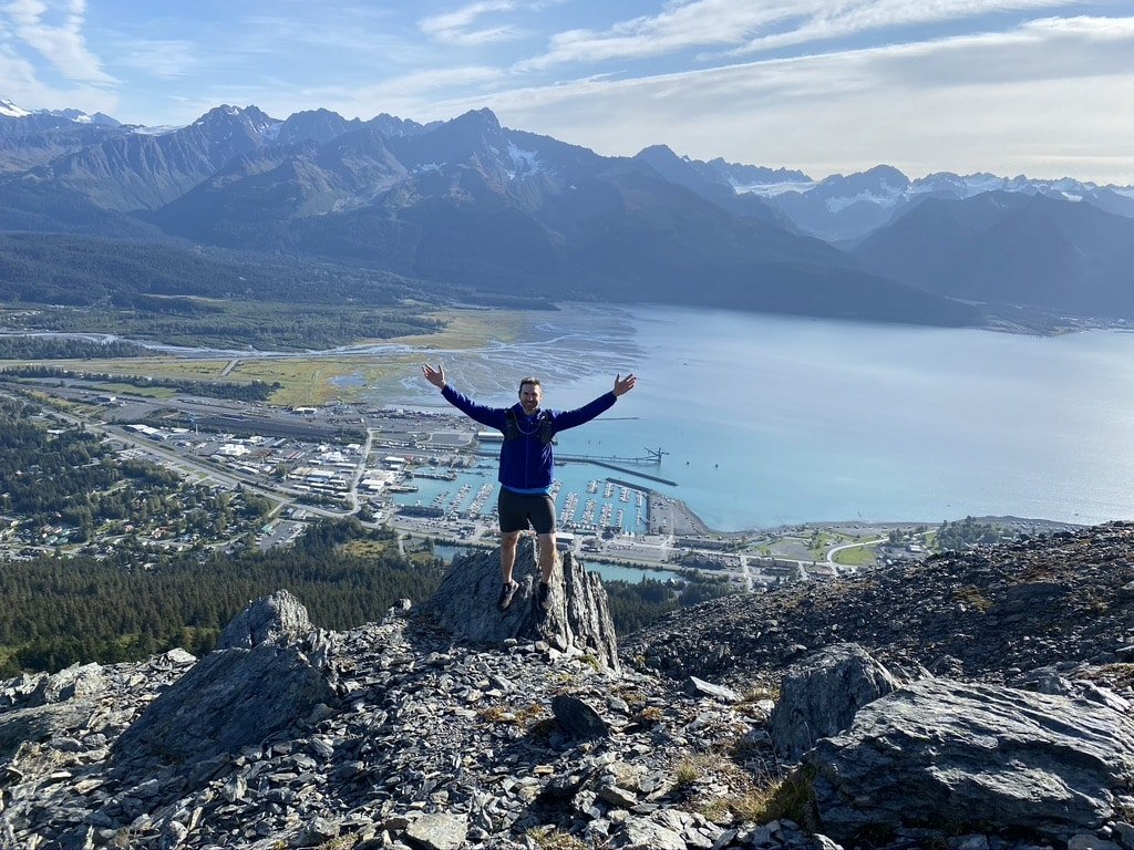 A man stands proudly with his arms raised on the edge of an overlook with Curry Ridge mountains and a bay full of boats laid out behind him