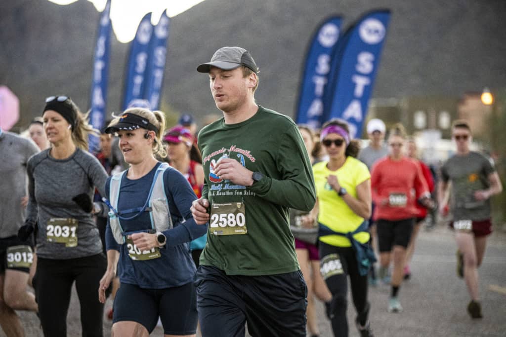 A wave of runners in running hats and hydration vests glide past the start line flags