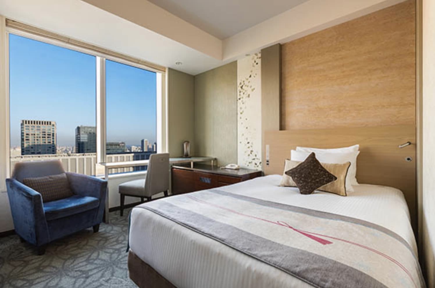 Queen bed in a sunny modern hotel room with large windows overlooking the city