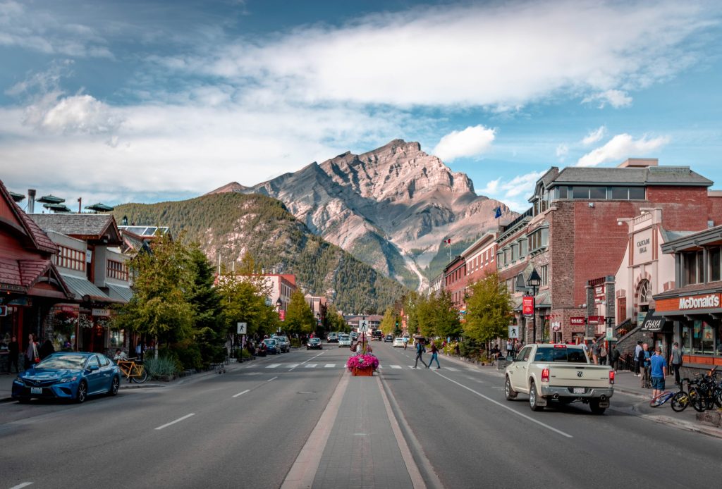 The main road of downtown Banff shows a quaint mountain town with mountains and trees rising above the buildings